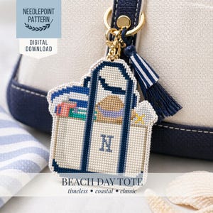 Könnte beinhalten: Ein Kreuzstichmuster für eine Strandtasche. Der Taschenanhänger ist weiß mit blauen Akzenten, einem goldfarbenen Verschluss und einer blau-weißen Quaste. Die Tasche ist mit Strandutensilien gefüllt und trägt den Buchstaben "N". Der Text lautet "Beach Day Tote".