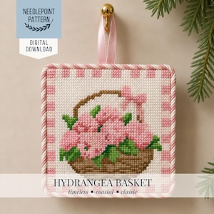 Op de afbeelding: Vierkantig kruissteekpatroon met een mandje roze hortensia's. Het ontwerp heeft een roze en wit geblokte rand en een gedraaide touwrand. De tekst "Hydrangea Basket" staat onderaan, met "tijdloos, kust, klassiek" eronder.