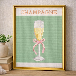 Op de afbeelding: Ingeklemd kruissteekkunstwerk met een champagneglas met een roze strik op een groene achtergrond. Het woord "CHAMPAGNE" is bovenaan in oranje gestikt. Het kunstwerk zit in een gouden lijst.