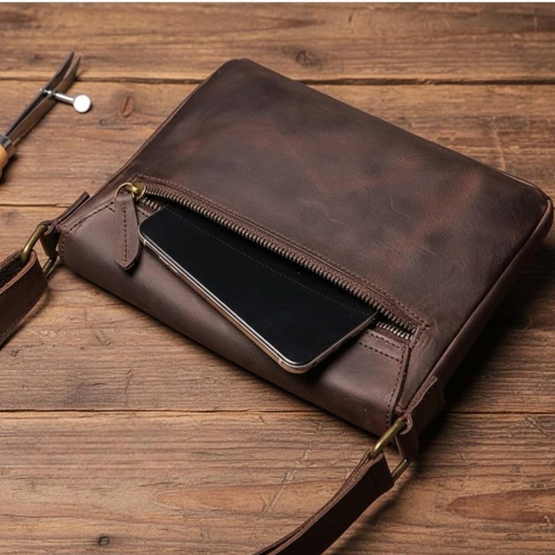 Peut inclure: Un sac bandouli&egrave;re en cuir marron avec une fermeture &eacute;clair, une longue bandouli&egrave;re r&eacute;glable et une tablette &agrave; l'int&eacute;rieur. Le sac est en cuir vieilli et a un aspect vintage. Le sac est sur une surface en bois.