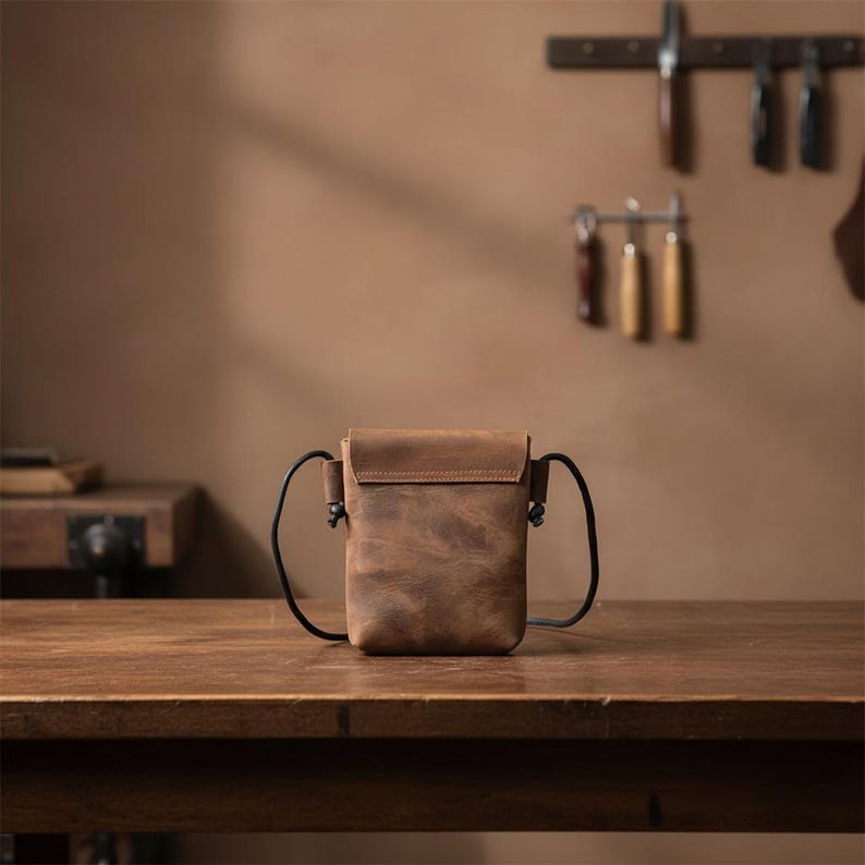 May include: A small, rectangular brown leather crossbody bag with a smooth flap closure and thin black cord straps attached to the sides. The leather has a matte, distressed texture with visible natural variations in the brown color throughout.