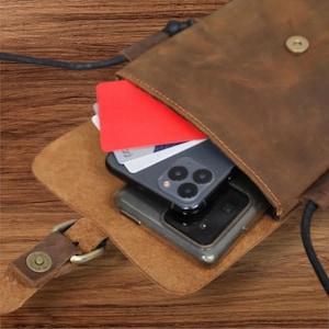May include: A brown leather crossbody bag with a fold-over flap secured by a metal snap button. The bag contains a dark gray smartphone with a triple-lens camera and several rectangular cards, including one bright red card, tucked inside the main compartment.
