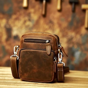 Puede incluir: Un bolso bandolera de cuero marrón con forma rectangular, que presenta un bolsillo frontal con cremallera y un compartimento principal con cierre de cremallera. Incluye una correa extraíble con ganchos metálicos en los anillos laterales.