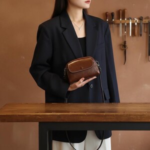 Pode incluir: Uma bolsa transversal de couro marrom com formato arredondado e fecho de aba. A bolsa tem uma al&ccedil;a de corrente prateada e uma borda costurada branca. A bolsa &eacute; segurada em frente a uma pessoa vestindo um blazer preto.