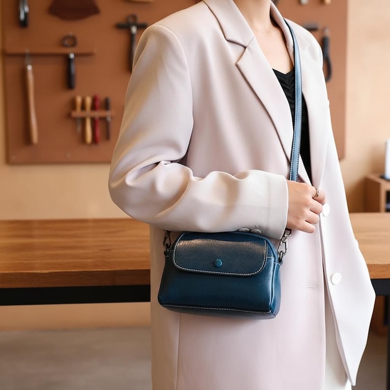 Pode incluir: Uma bolsa transversal de couro azul-petr&oacute;leo com fecho de aba e detalhe de bot&atilde;o. A bolsa tem uma al&ccedil;a fina e ajust&aacute;vel e detalhes de corrente prateada. A bolsa &eacute; usada sobre um blazer rosa claro.