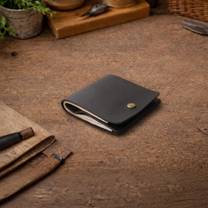 Puede incluir: Cartera de cuero marrón oscuro con cierre de botón a presión de latón. La cartera rectangular parece estar llena de papeles doblados. El fondo es una superficie de madera.