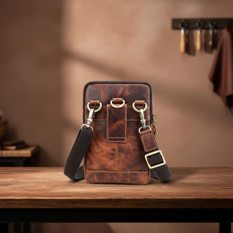 May include: A brown leather crossbody bag with a distressed finish. The bag has a rectangular shape with a zipper closure and adjustable black strap. Features silver-tone hardware and a small belt loop on the back. The bag is sitting on a wooden surface.