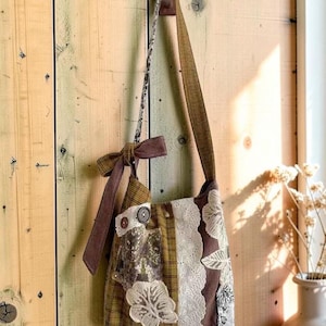 Op de afbeelding: Een patchwork schoudertas gemaakt van bruine, olijfgroene en crèmekleurige stoffen. De tas is voorzien van een lange schouderband, een decoratieve strik aan de zijkant, kanten afwerking en bloemenborduursels. Het is gemaakt van diverse texturen.