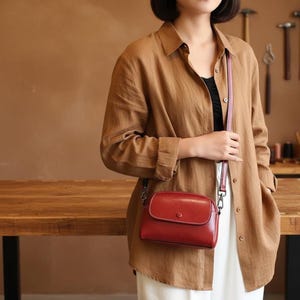Pode incluir: Uma bolsa transversal de couro vermelho com fecho de aba e detalhe de bot&atilde;o. A bolsa tem uma al&ccedil;a fina rosa e ferragens prateadas. A bolsa &eacute; usada sobre uma camisa marrom e cal&ccedil;as brancas.