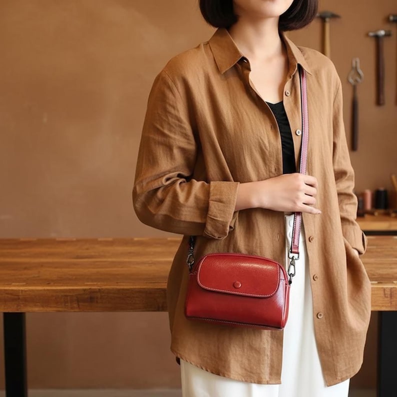 Pode incluir: Uma bolsa transversal de couro vermelho com fecho de aba e detalhe de bot&atilde;o. A bolsa tem uma al&ccedil;a fina rosa e ferragens prateadas. A bolsa &eacute; usada sobre uma camisa marrom e cal&ccedil;as brancas.