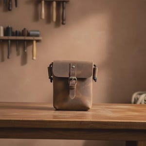 以下が含まれることがあります： フラップとバックル留めの茶色の革製クロスボディバッグ。長方形のバッグは木製のテーブルの上にあります。背景はニュートラルカラーで、壁にはツールが吊り下げられています。