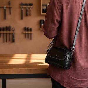 Pode incluir: Uma bolsa transversal de couro preto com formato arredondado e uma al&ccedil;a longa e ajust&aacute;vel. A bolsa apresenta costuras brancas e um pequeno fecho de aba. A pessoa est&aacute; vestindo uma camisa marrom.
