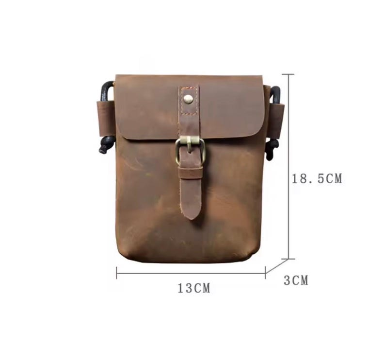 May include: A brown leather crossbody bag with a front flap, a metal buckle closure, and black side cord attachments. The bag measures 18.5 cm in height, 13 cm in width, and 3 cm in depth as indicated by the visible measurement lines.
