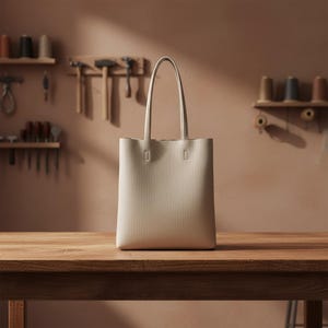 Puede incluir: Un bolso tote beige con asas largas se encuentra sobre una mesa de madera. El bolso tiene forma rectangular y un diseño minimalista. El fondo muestra un taller con herramientas y estanterías.