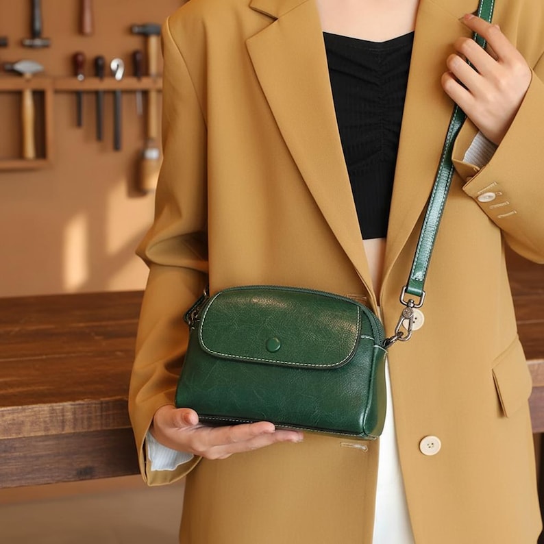 Pode incluir: Uma bolsa transversal de couro verde escuro com fecho de aba e costura branca. A bolsa tem uma al&ccedil;a verde combinando e ferragens prateadas. A pessoa est&aacute; usando um blazer bege e um top preto.