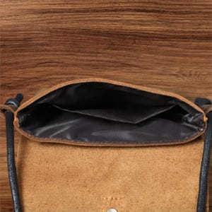 May include: A tan suede crossbody bag with a black interior lining and a black cord strap. The bag features an open top compartment with a single internal pocket and a visible metal snap button closure on the front flap exterior.