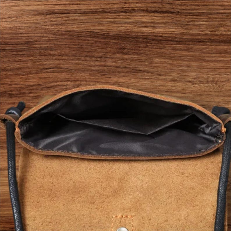 May include: A tan suede crossbody bag with a black interior lining and a black cord strap. The bag features an open top compartment with a single internal pocket and a visible metal snap button closure on the front flap exterior.