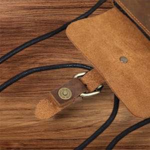 May include: A brown leather pouch featuring a fold-over flap with a single metal snap button closure. The pouch is attached to a thin black cord strap that loops through a metal ring connector, secured by a small leather tab with a metal rivet.