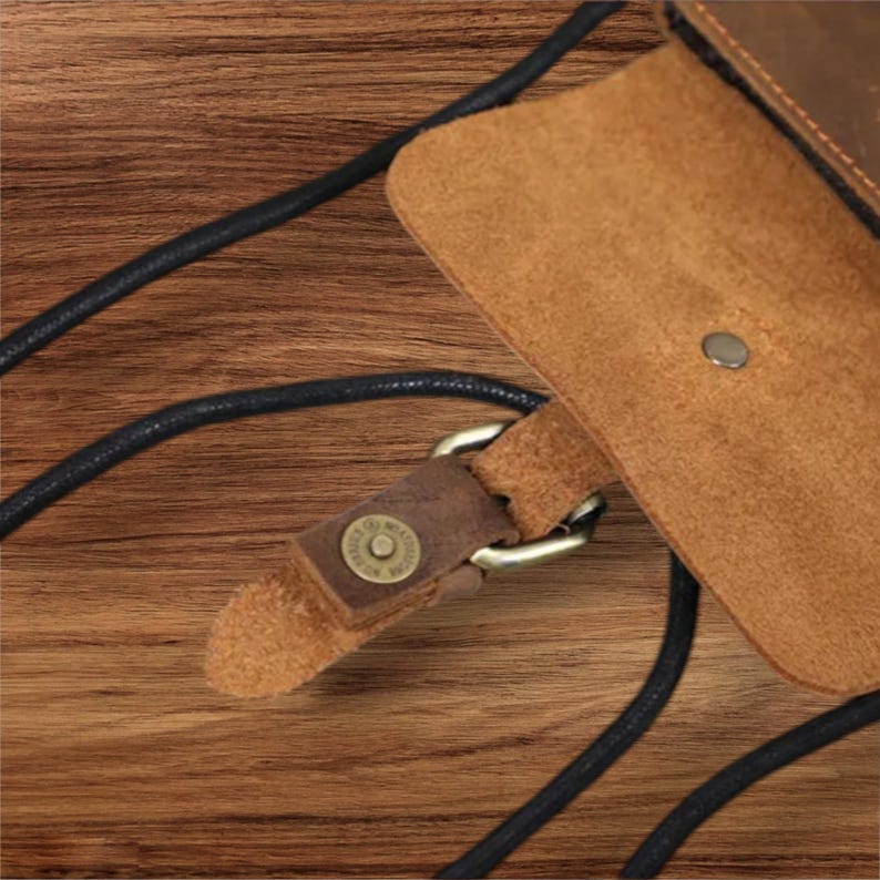 May include: A brown leather pouch featuring a fold-over flap with a single metal snap button closure. The pouch is attached to a thin black cord strap that loops through a metal ring connector, secured by a small leather tab with a metal rivet.