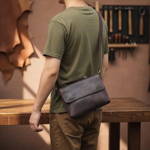 Peut inclure: Un sac messager en cuir marron fonc&eacute; avec une fermeture &agrave; rabat est port&eacute; en bandouli&egrave;re. Le sac a une longue et fine bandouli&egrave;re et est port&eacute; par une personne portant un t-shirt vert et un pantalon marron. L'arri&egrave;re-plan montre une table en bois et des outils.