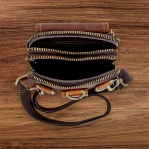 May include: Brown leather crossbody bag with multiple zippered compartments. The bag features a brown canvas strap, bronze-colored hardware, and a top flap. The bag is open, revealing the interior compartments.