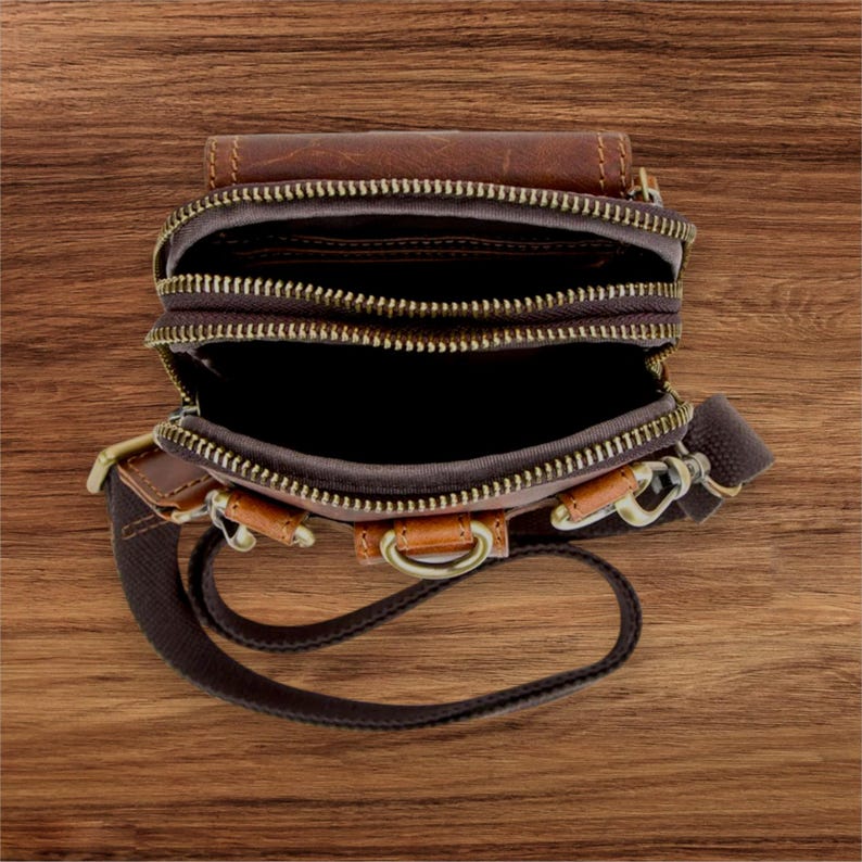 May include: Brown leather crossbody bag with multiple zippered compartments. The bag features a brown canvas strap, bronze-colored hardware, and a top flap. The bag is open, revealing the interior compartments.