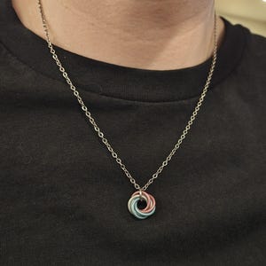 May include: A silver chain necklace with a pendant featuring a circular design of interlocking rings in pastel blue, pink, and white. The necklace is worn against a black top.