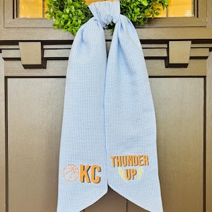 May include: A blue and white gingham door scarf with the text "KC" and "THUNDER UP" in orange and yellow. The scarf is tied at the top and hangs down. A green wreath is visible in the background.