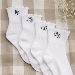May include: Four white ankle socks with ruffled tops, each embroidered with a different set of initials in black thread. The socks are arranged on a checkered beige and white surface, with a sprig of white flowers in the background.
