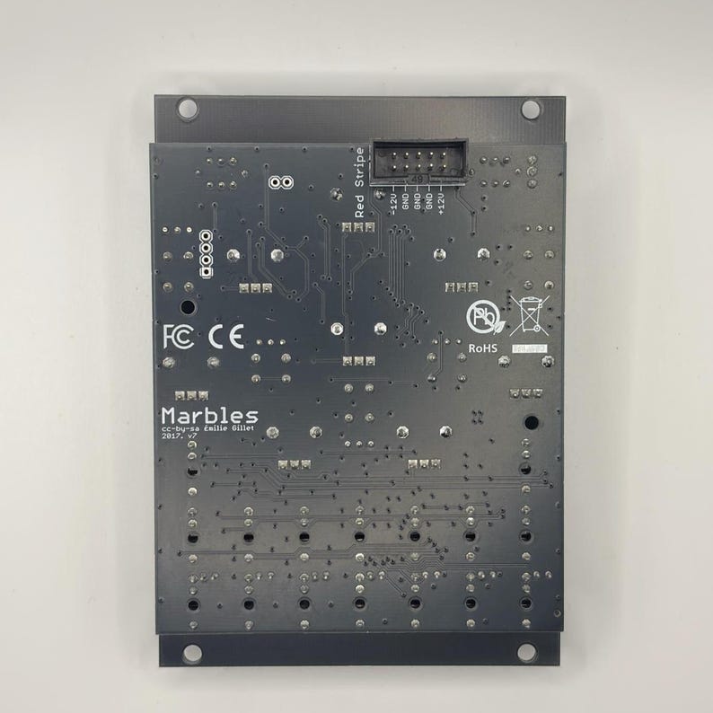 May include: A dark gray circuit board with the text "Marbles" and "Red Stripe". The board has various electronic components, connectors, and the RoHS symbol. The board is rectangular with a metal frame.