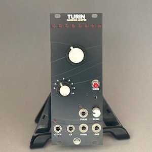 Può includere: Un dispositivo elettronico nero con l'etichetta "TURIN RANDOM LOOPER" con più manopole, pulsanti e prese di ingresso/uscita. Il dispositivo ha una serie di luci rosse e testo bianco. È montato su un supporto nero.