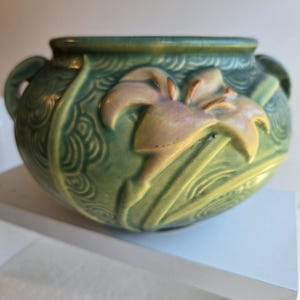 May include: A green ceramic bowl with a floral design. The bowl has two small handles and a raised iris flower in shades of cream and yellow. The background has a textured, swirling pattern.