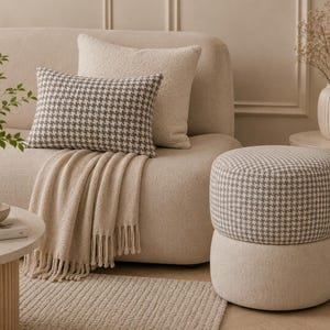 Può includere: Un divano beige con un cuscino e un pouf a fantasia pied-de-poule. Una coperta beige con frange è drappeggiata sul divano. Un tavolino rotondo contiene una ciotola e un libro. Un vaso con fiori secchi è su un tavolino.