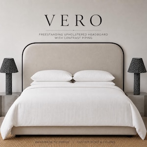 Op de afbeelding: Een vrijstaand gestoffeerd hoofdbord met het label VERO en contrasterende biezen. Het hoofdbord is beige met een donkerbruine rand, geplaatst achter een bed met wit beddengoed en twee zwarte lampen met patronen op bijzettafels.