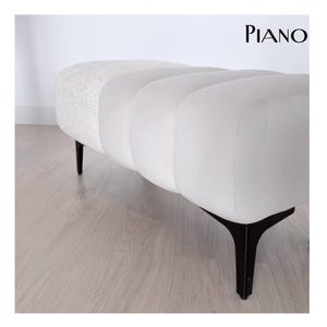 Pode incluir: Um banco estofado branco com uma seção final texturizada e um design canelado. O banco tem pernas escuras e anguladas e a palavra "PIANO" em texto preto no canto superior direito. O banco tem aproximadamente 152 cm de comprimento.
