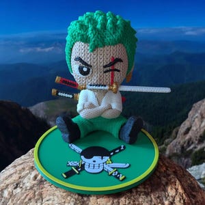 Puede incluir: Figura de ganchillo de un personaje con pelo verde, camisa blanca y pantalón verde, sosteniendo una espada en la boca. La figura está sentada sobre una base circular verde con un diseño de calavera y huesos cruzados. El fondo muestra una cordillera.