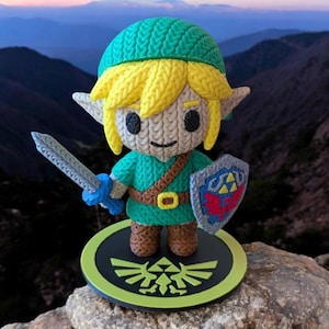 Könnte beinhalten: Handgefertigte, gestrickte Figur einer Figur aus The Legend of Zelda. Die Figur trägt eine grüne Tunika, einen grünen Hut und hat blondes Haar. Sie hält ein Schwert und einen Schild mit einem Triforce-Symbol. Die Figur steht auf einem schwarz-grünen Sockel.