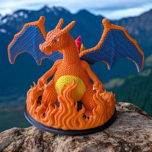 May include: An orange and blue Charizard figurine, with flames at its base, stands on a rock. The dragon has blue wings, a yellow belly, and a knitted texture. The background features a blurred mountain range.
