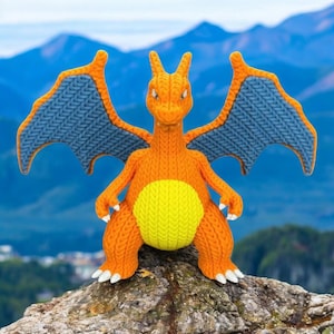 Könnte beinhalten: Eine gestrickte Charizard-Figur in Orange und Blau mit gelben Akzenten. Die Figur ist auf einem Felsen platziert, die Flügel sind ausgebreitet. Der Hintergrund zeigt verschwommene Berge und einen blauen Himmel.