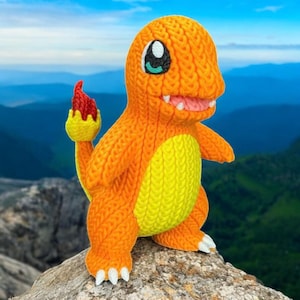 May include: A knitted orange Charmander figure with a yellow belly and a red flame on its tail. The figure has white teeth and a smiling expression. It is posed on a rock with a mountain backdrop.