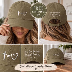 May include: An olive green baseball cap featuring white embroidery on the front with a cross symbol, an equals sign, and a heart. The back of the cap displays the white embroidered text, "We love because He first loved us. 1 JOHN 4:19."
