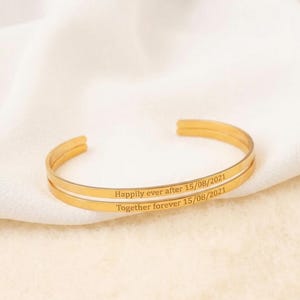 Puede incluir: Una pulsera de brazalete dorada con el texto grabado "Happily ever after 15/08/2021" y "Together forever 15/08/2021". La pulsera se muestra sobre una superficie blanca texturizada.