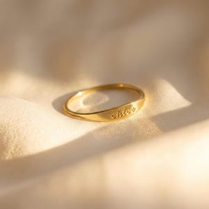 Könnte beinhalten: Ein goldfarbener Ring mit dem eingravierten Namen "chloe" auf der Vorderseite. Der Ring liegt auf einem weichen, cremefarbenen Stoff. Der Ring hat ein schlichtes Banddesign.