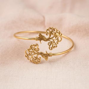 Peut inclure: Un bracelet jonc ouvert doré orné de deux motifs d'arbre en filigrane symétriques et complexes à chaque extrémité. La tige métallique est lisse et circulaire, se connectant aux détails en forme de branches tourbillonnantes.