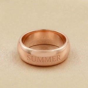 Peut inclure: Une bague en or rose avec le mot "SUMMER" gravé sur le bandeau extérieur. La bague a une finition lisse et polie et une forme arrondie. La bague est un bijou.