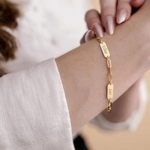 Op de afbeelding: Goudkleurige schakelarmband met twee rechthoekige naamplaatjes. Eén plaatje leest "Eliza" en de andere "Leo". De armband wordt om een pols gedragen, met een wit overhemd op de achtergrond. Een delicaat sieraad.