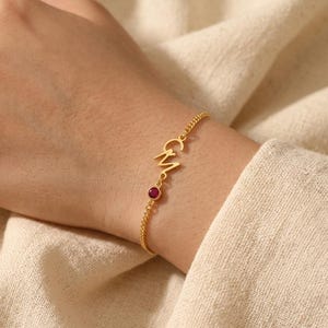 Puede incluir: Una pulsera dorada con las letras "GM" y una pequeña gema roja redonda. La pulsera está en una cadena fina. La pulsera se lleva en la muñeca.