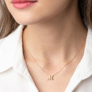Puede incluir: Un collar dorado con una fina cadena y un colgante con la letra "M" en cursiva. El collar se lleva sobre una camisa blanca. El colgante está centrado en el pecho.