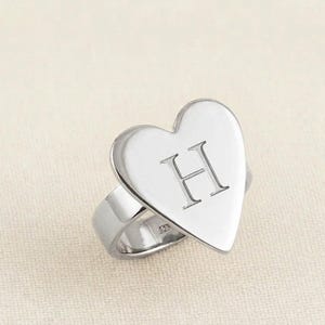 May include: A silver ring featuring a heart-shaped face with the letter "H" engraved. The ring is set against a plain background. The ring is made of a shiny, reflective metal.