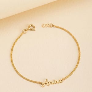 Könnte beinhalten: Ein zartes goldenes Armband mit dem Namen "Amina" in Schreibschrift. Das Armband hat ein feines Kettendesign und einen sicheren Verschluss. Der Schmuck wird vor einem hellbeigen Hintergrund präsentiert.